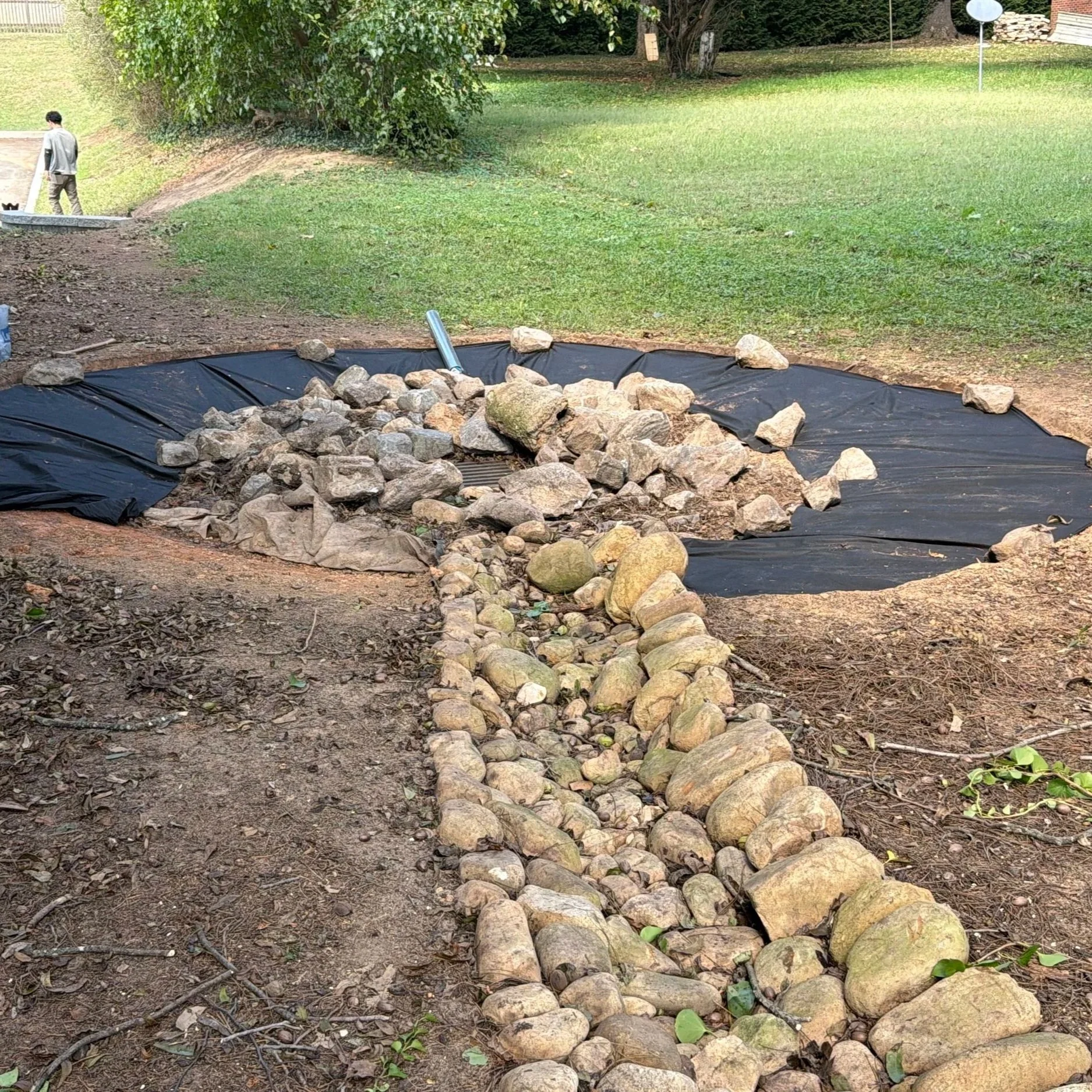 Backyard dry creek bed under construction with landscaping fabric, rocks, and a worker nearby