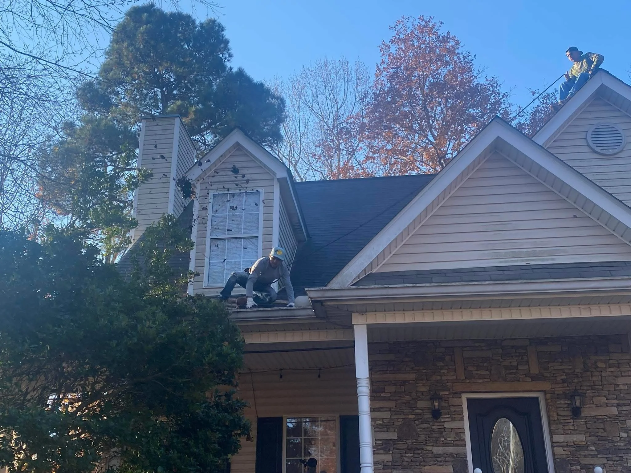 Two workers repairing the roof of a suburban house, with one person on the roof peak and another crouching near the eaves.
