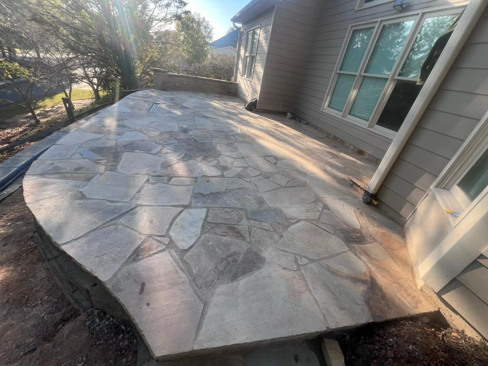 Flagstone patio beside a gray-sided house with windows and a low stone retaining wall