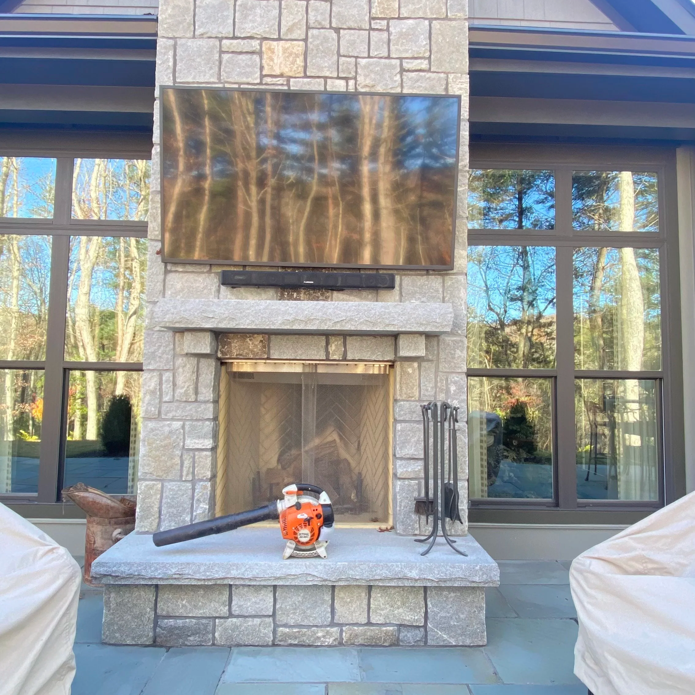 Stone fireplace with mounted TV, soundbar, fireplace tools, and orange leaf blower on the hearth