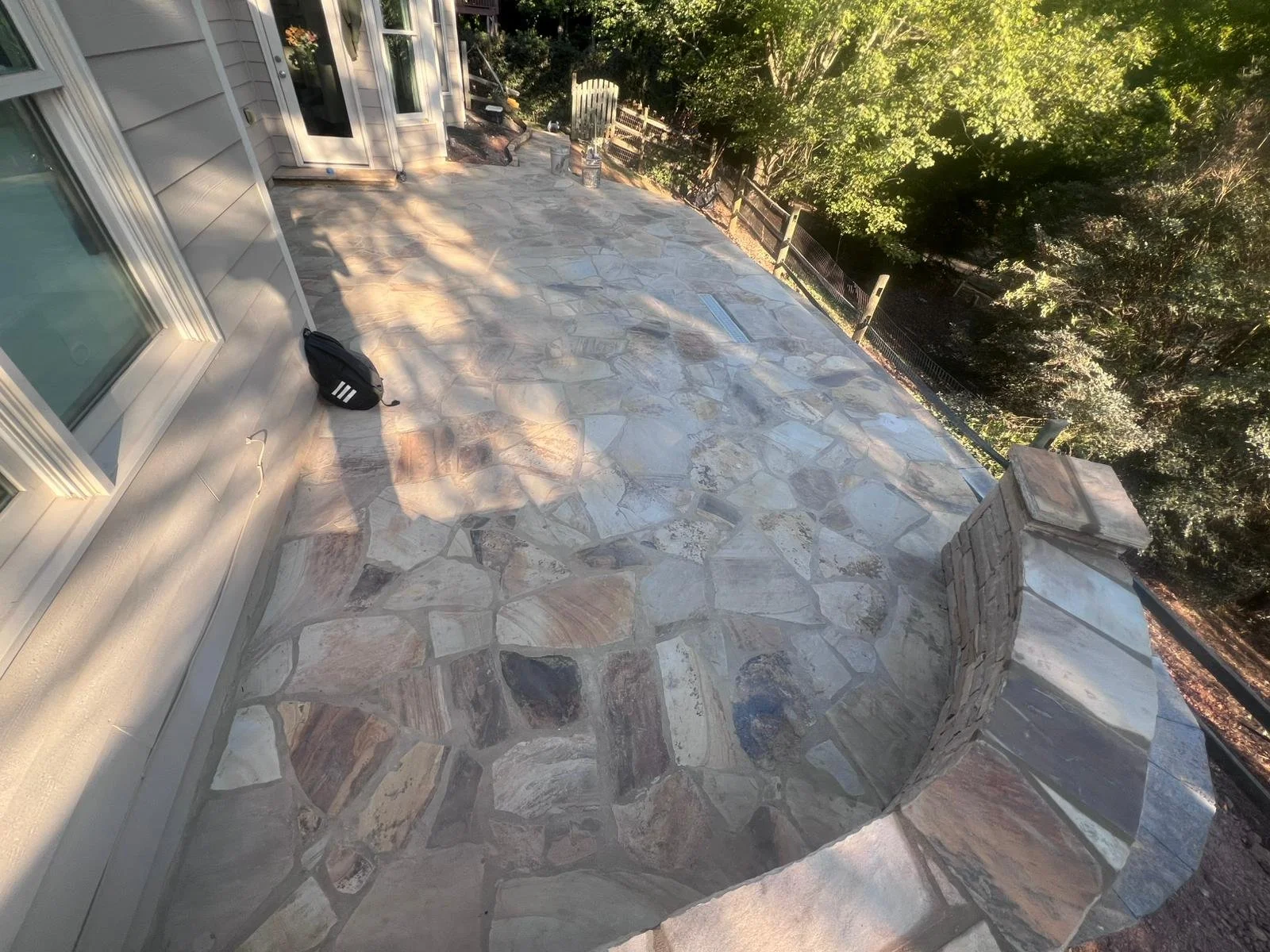 Flagstone patio beside a house, bordered by a wooded yard and railing in sunlight