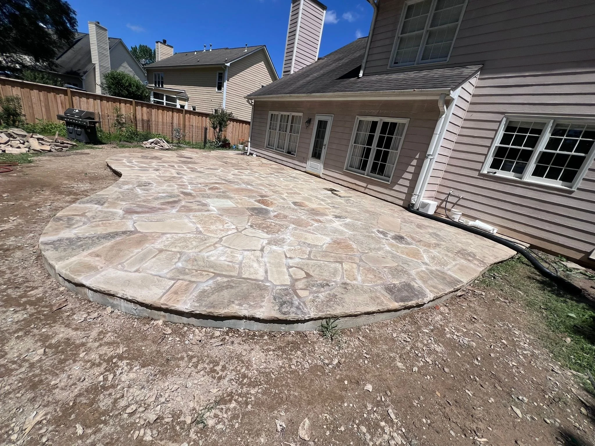 Large irregular flagstone patio under construction beside a suburban house, with a backyard grill and wooden privacy fence.