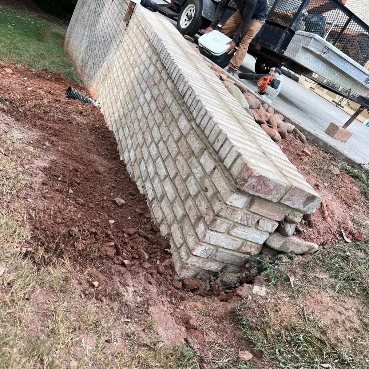 Damaged stone retaining wall with eroded soil, drainage pipe, landscaping rocks, and landscaping equipment trailer nearby