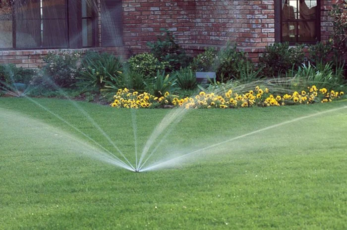 Lawn sprinkler watering a green lawn in front of a brick house and flower garden