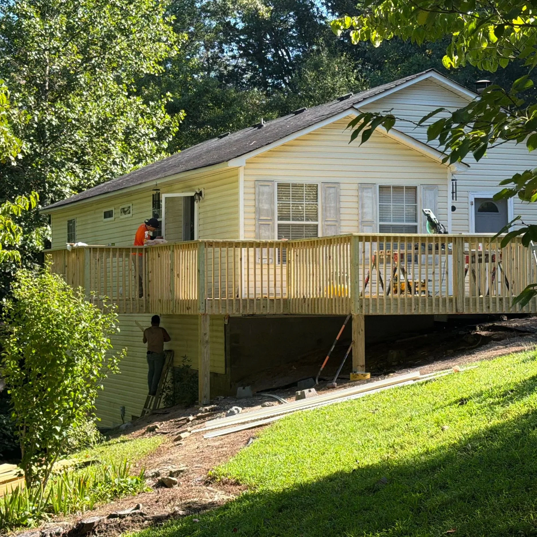 Workers renovating a raised house with a new wooden deck in a tree-lined yard