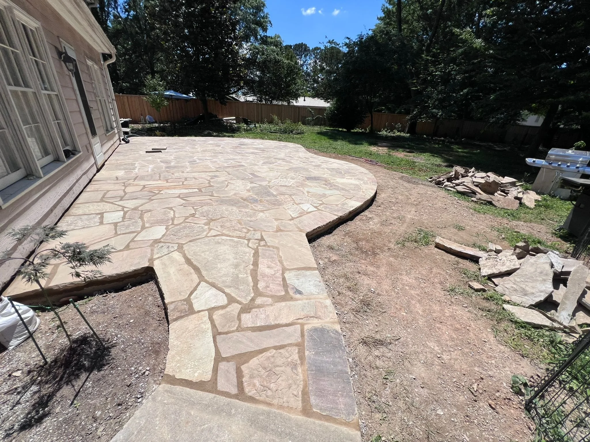 Backyard with a large natural stone patio, garden area, wooden privacy fence, trees, and outdoor grill
