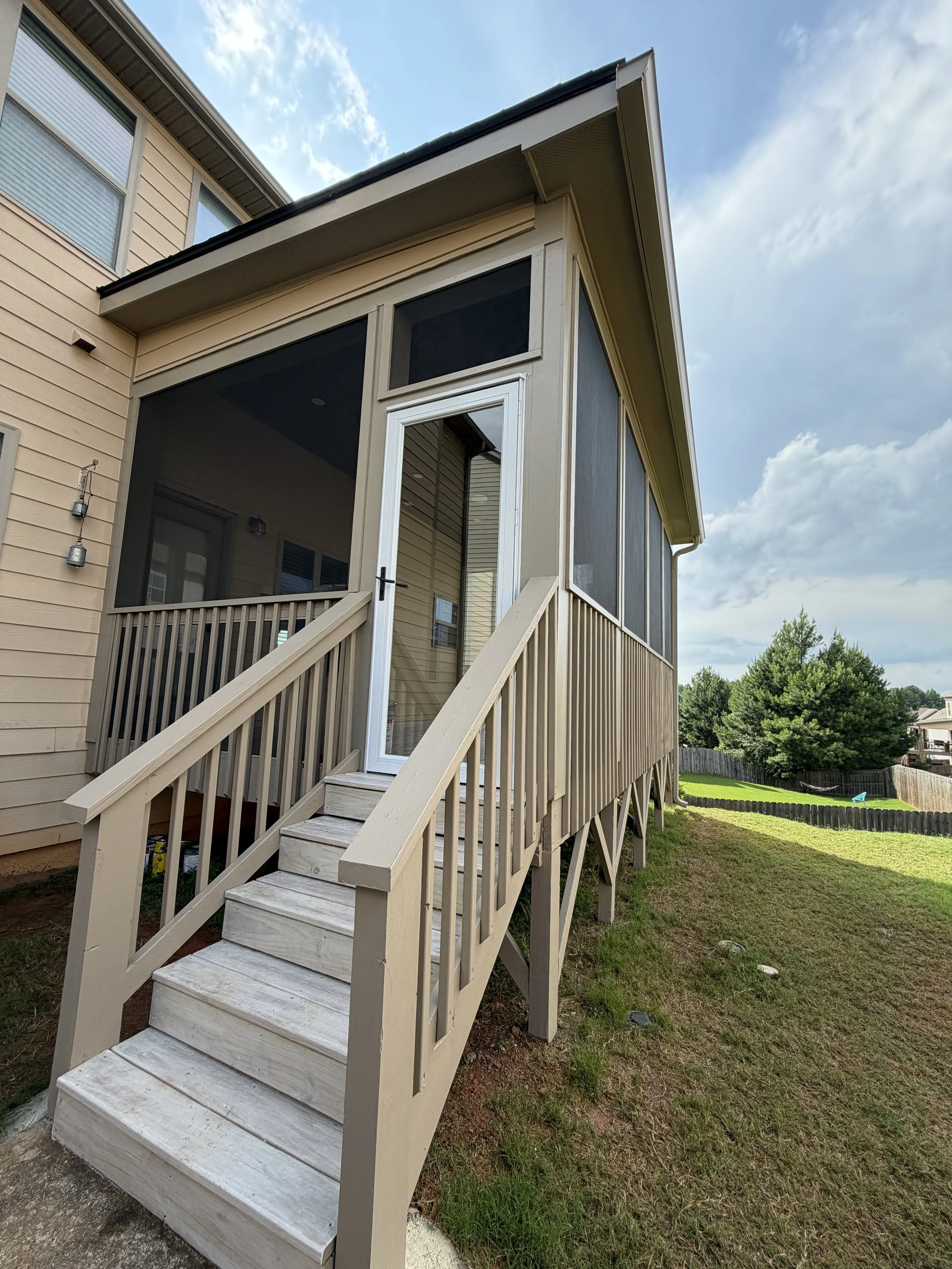 Exterior view of a tan-sided home with screened porch, wooden stairs and railing, backyard lawn, and blue sky.