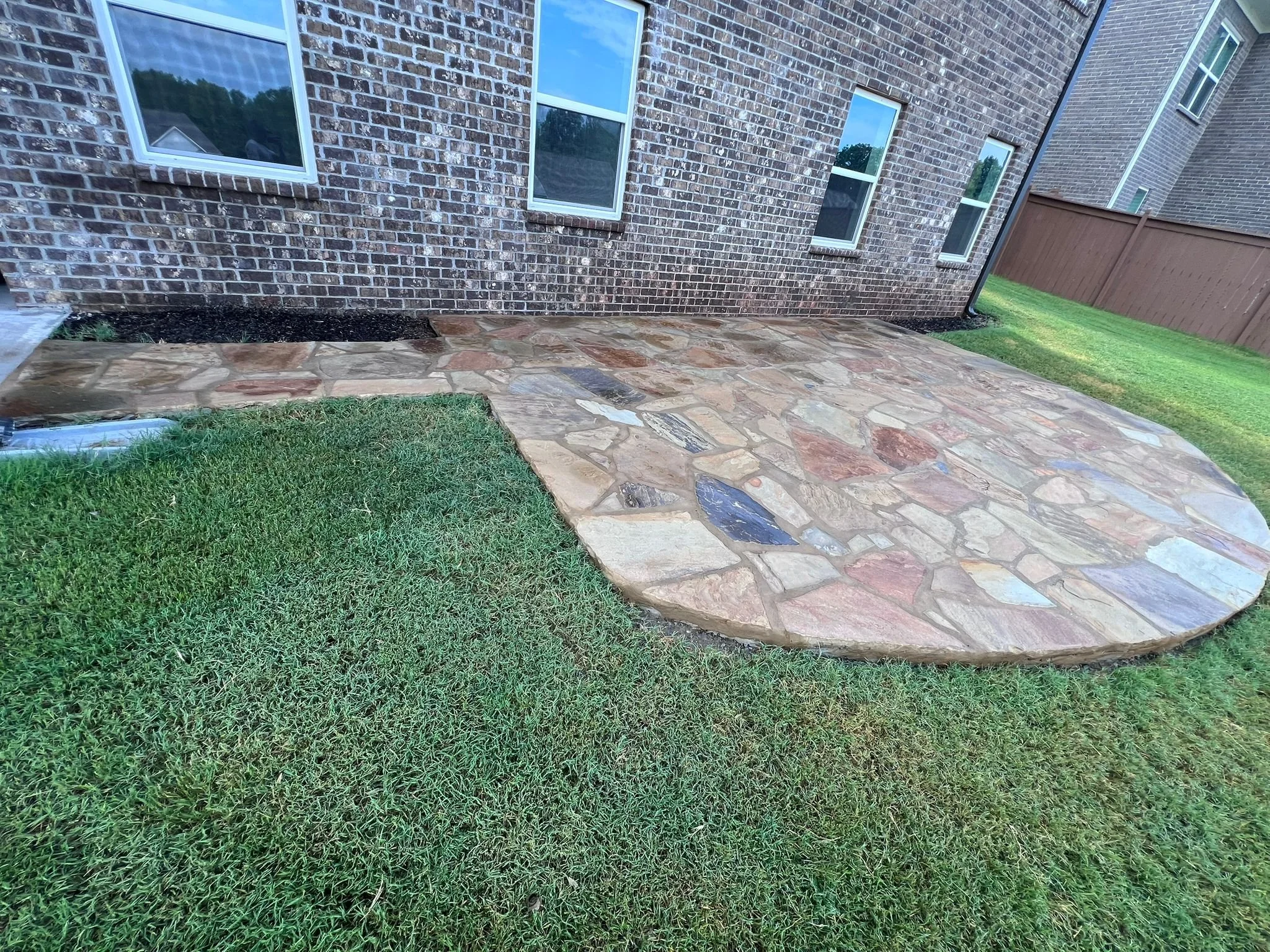 Irregular flagstone patio beside a brick house, bordered by green grass and a wooden fence.