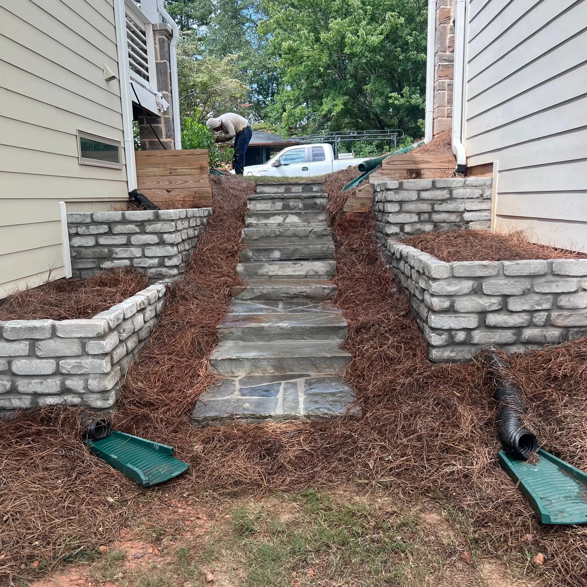Stone steps between two houses with landscaped retaining walls, drainage pipes, and a worker in the background