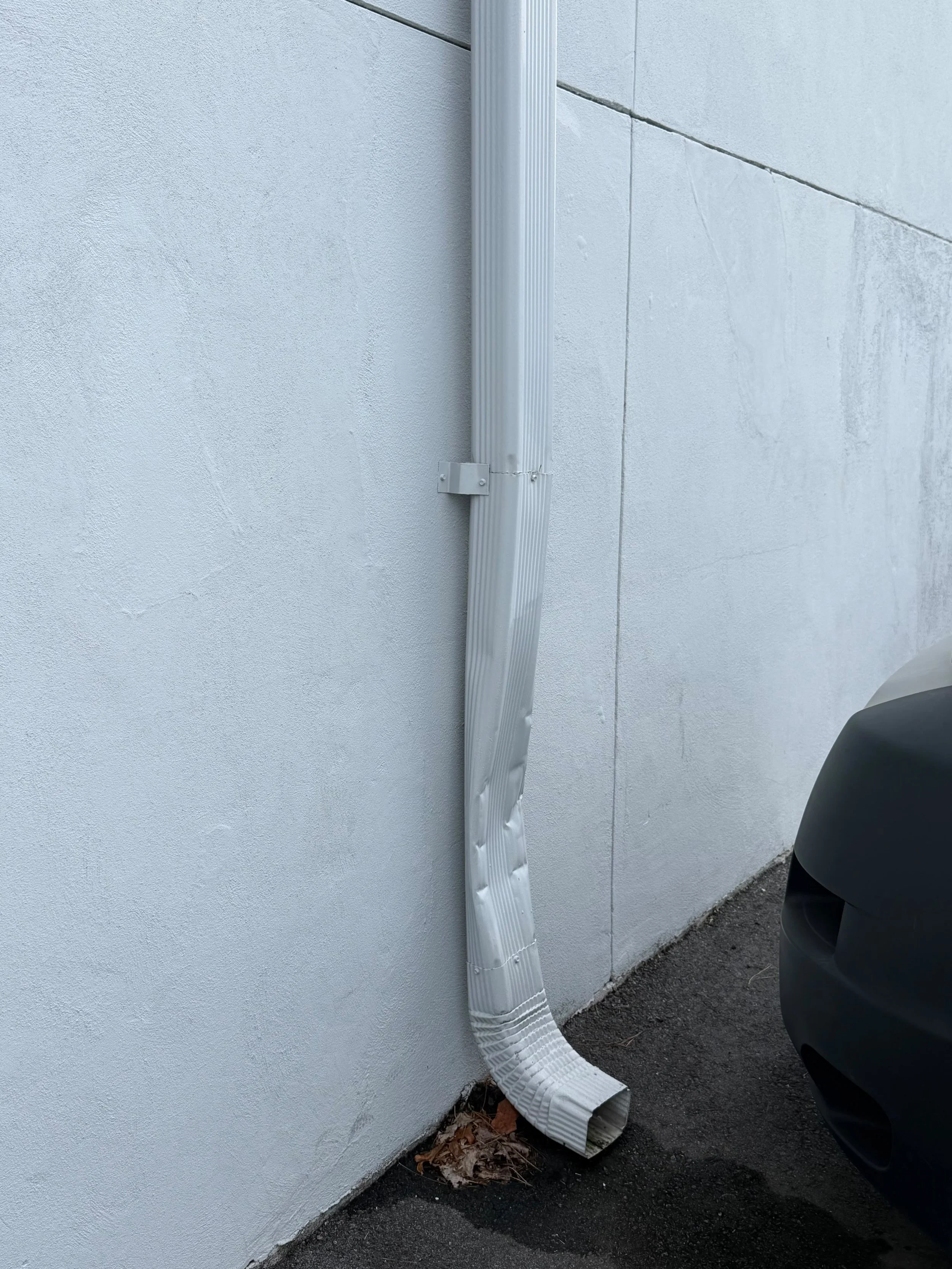 Damaged white downspout beside a building wall and parked car
