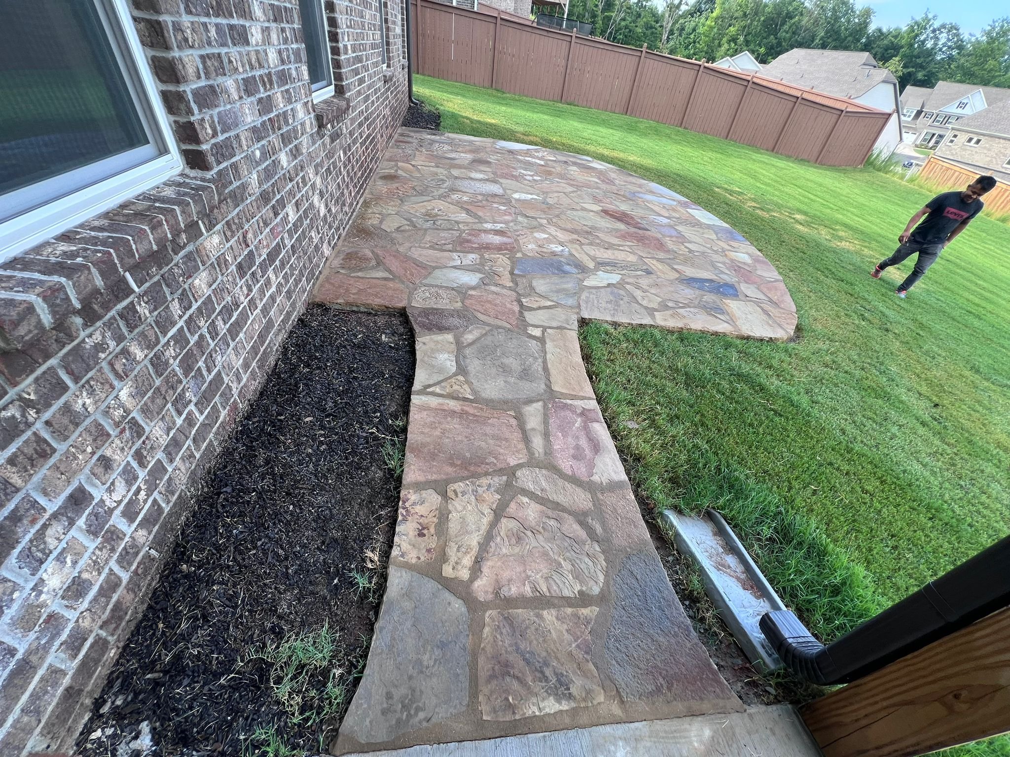 Flagstone patio and walkway beside a brick house, with a man standing on the lawn and fenced suburban homes in the background