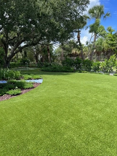 Lush landscaped backyard with manicured green lawn, tropical palm trees, mature shade trees, and garden beds under a sunny blue sky