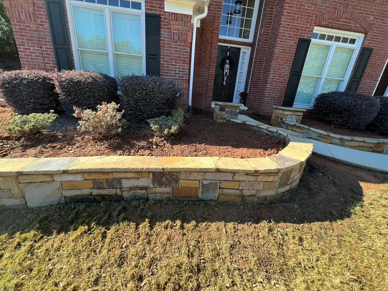 Brick house entrance with landscaped front garden, stone retaining walls, shrubs, and curved walkway