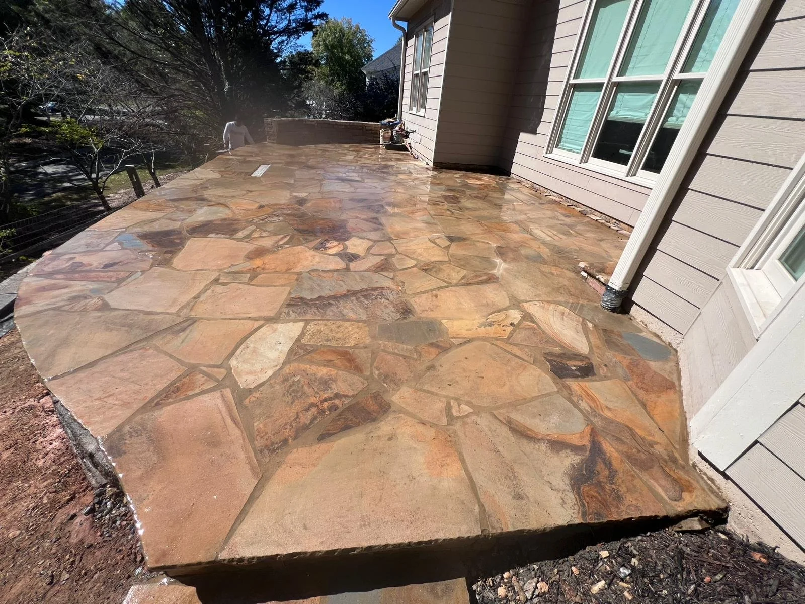 Large natural flagstone patio beside a beige-sided house, surrounded by landscaping and trees.