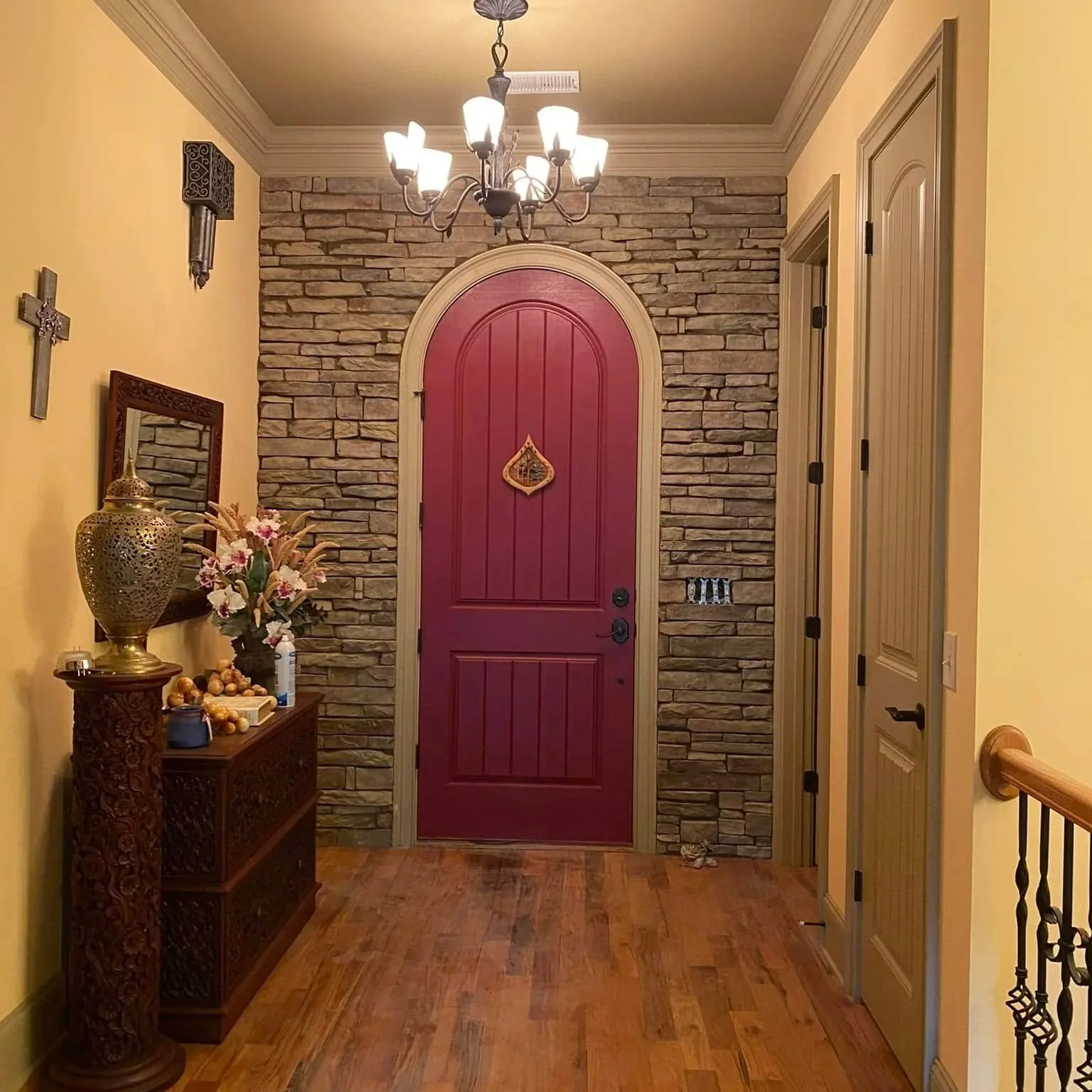 Elegant home entryway with a burgundy arched front door, stacked-stone accent wall, chandelier, decorative cabinet, floral arrangement, and staircase railing