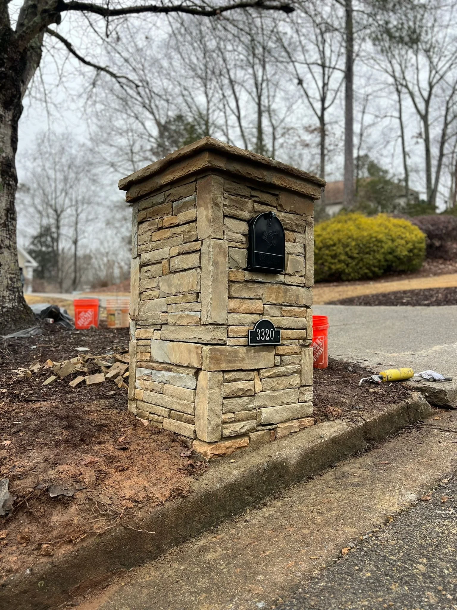 Stone-clad residential mailbox pillar with black mailbox and house number 3320 beside a wooded driveway