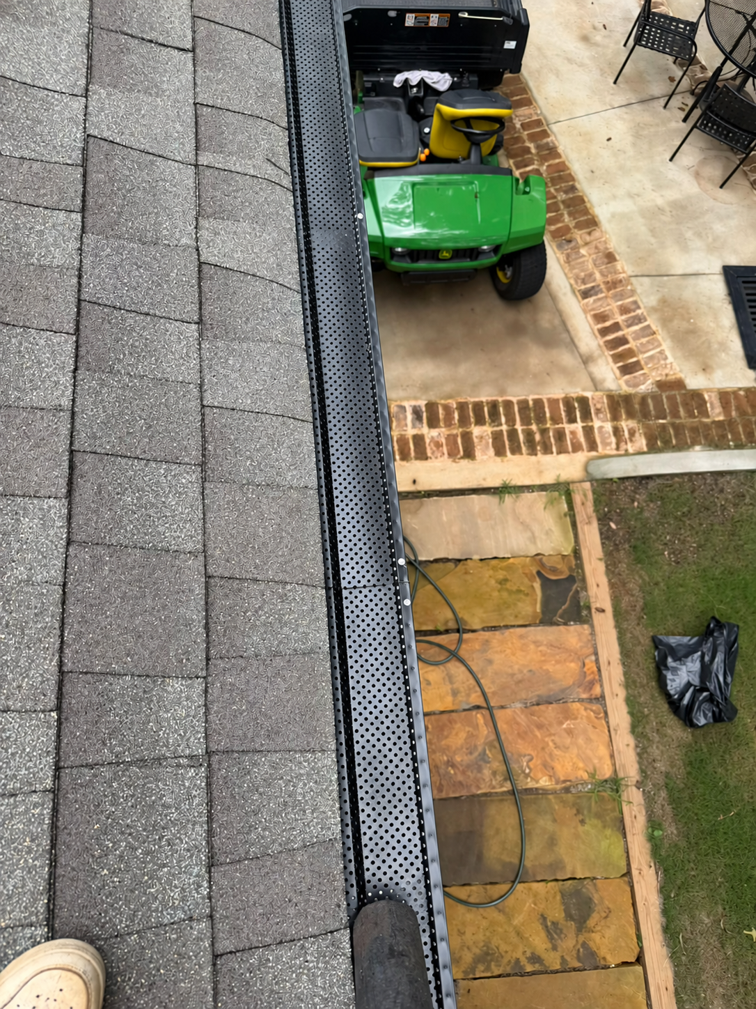 View from a shingle roof showing a black perforated gutter guard, green riding lawn mower, brick patio, and outdoor furniture below.
