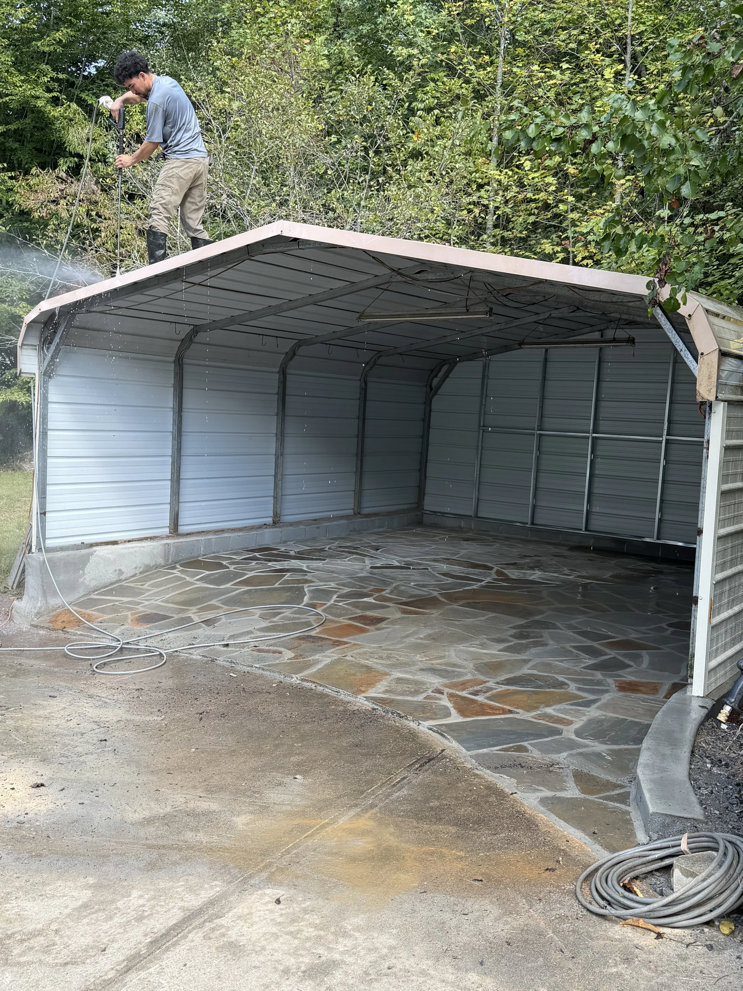 Man pressure-washing the roof of a metal carport surrounded by trees