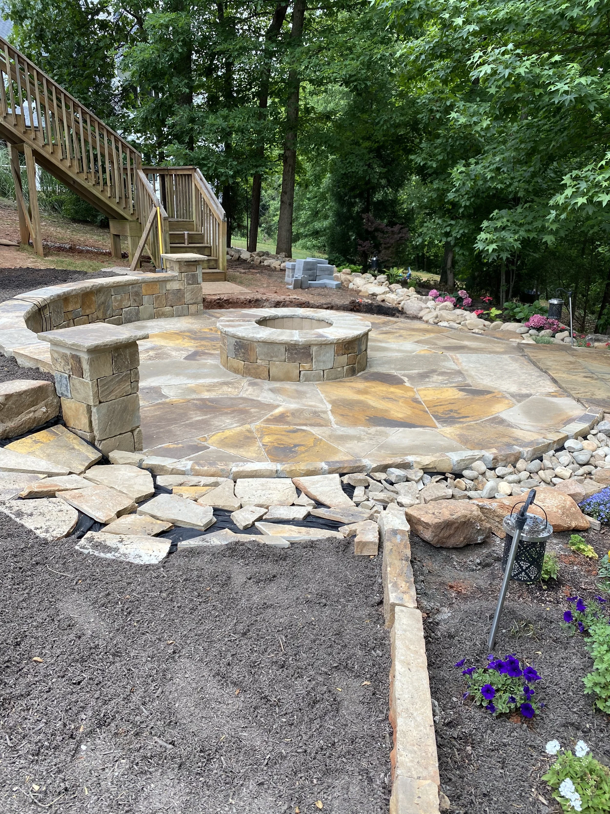 Landscaped backyard patio with a stone fire pit, wooden deck stairs, garden beds, flowers, and mature trees