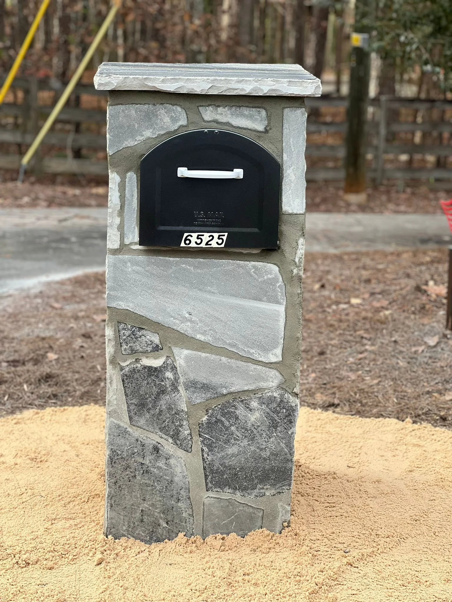 Black mailbox set in a stone-clad pillar, labeled 6525, beside a wooded driveway.