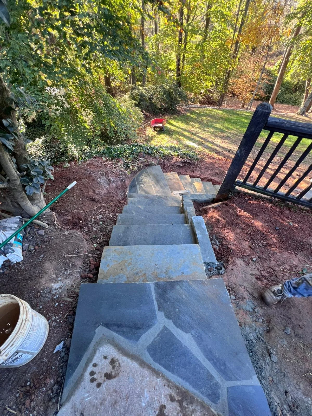 Stone steps with black railing descending through a wooded backyard toward a red wheelbarrow.