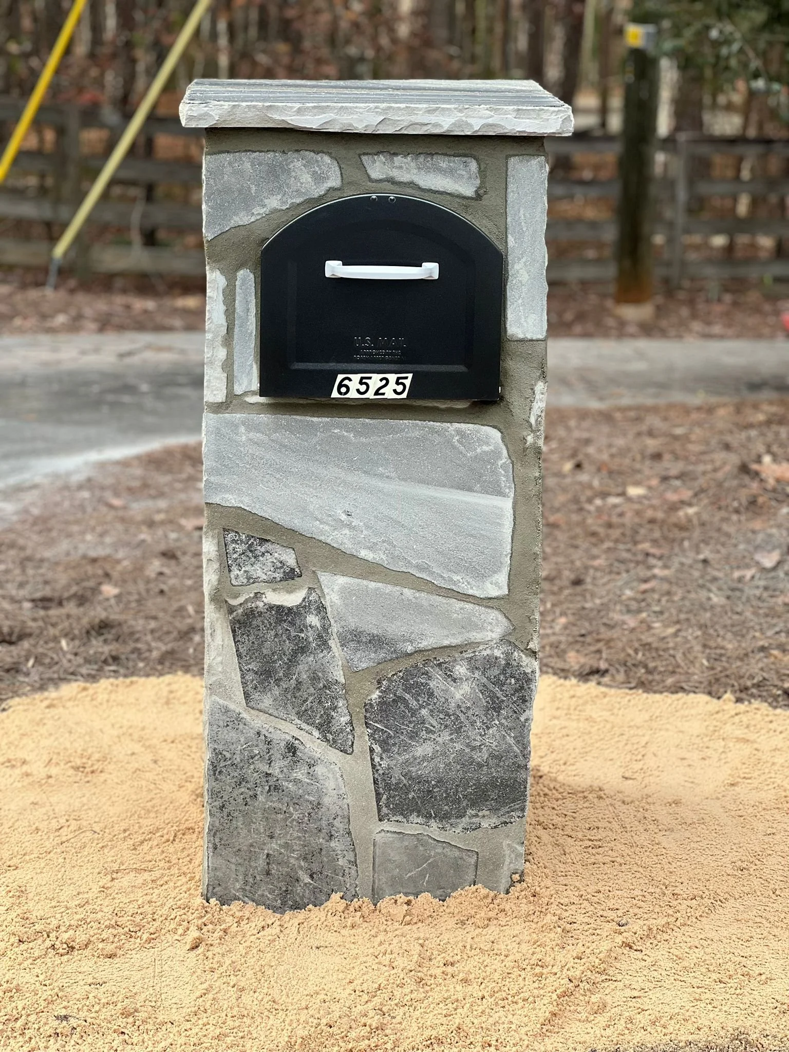 Black mailbox labeled 6525 mounted on a stone pillar beside a wooded driveway