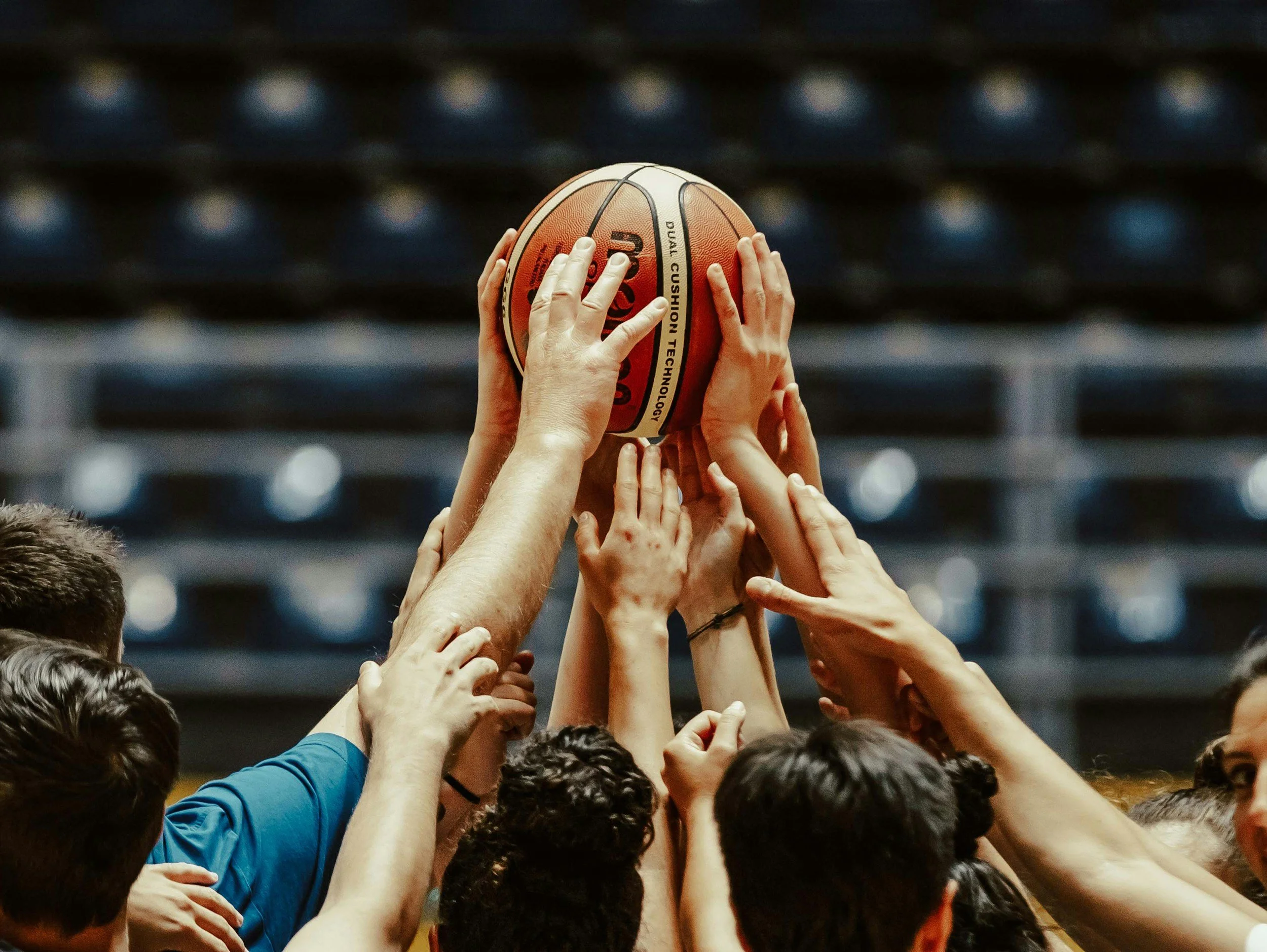Multiple hands reaching out to hold or support a basketball, with a blurred background of seating and lights, indicating a basketball game or practice.