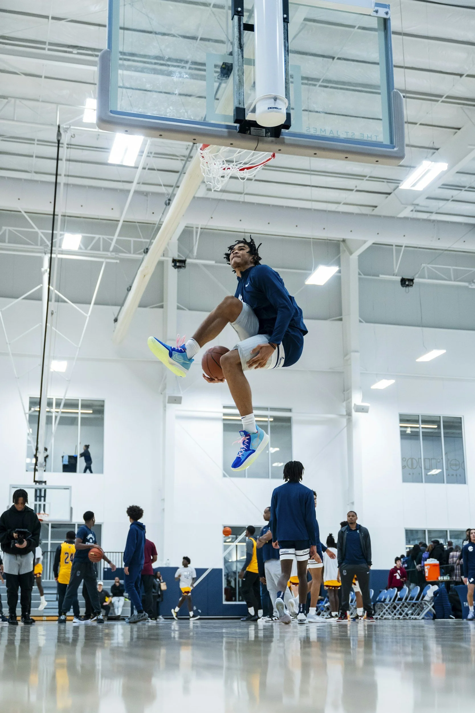 A basketball player mid-air dunking a basketball during practice in an indoor gym, with other players and coaches in the background.