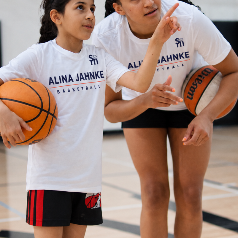 A young girl holding a basketball and a coach or trainer pointing, both wearing white 'Alina Jahnke Basketball' t-shirts on a gym basketball court.