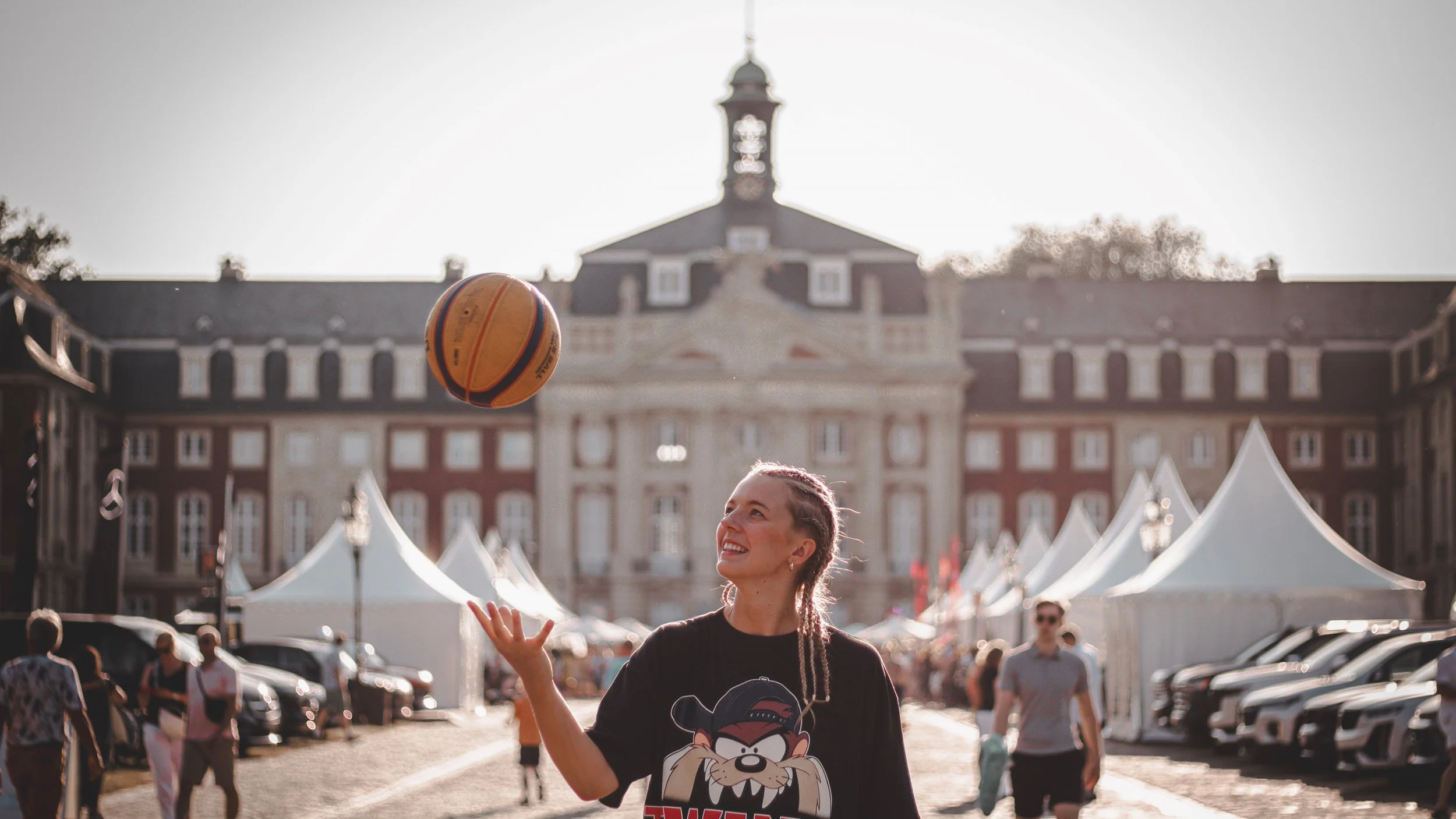 A young woman with braided hair is smiling and looking at a basketball that she has just tossed into the air. She is wearing a black T-shirt with a cartoon character from Looney Tunes. The scene is set outdoors in a busy area with people, tents, and parked cars, with a large historic building with a clock tower in the background.