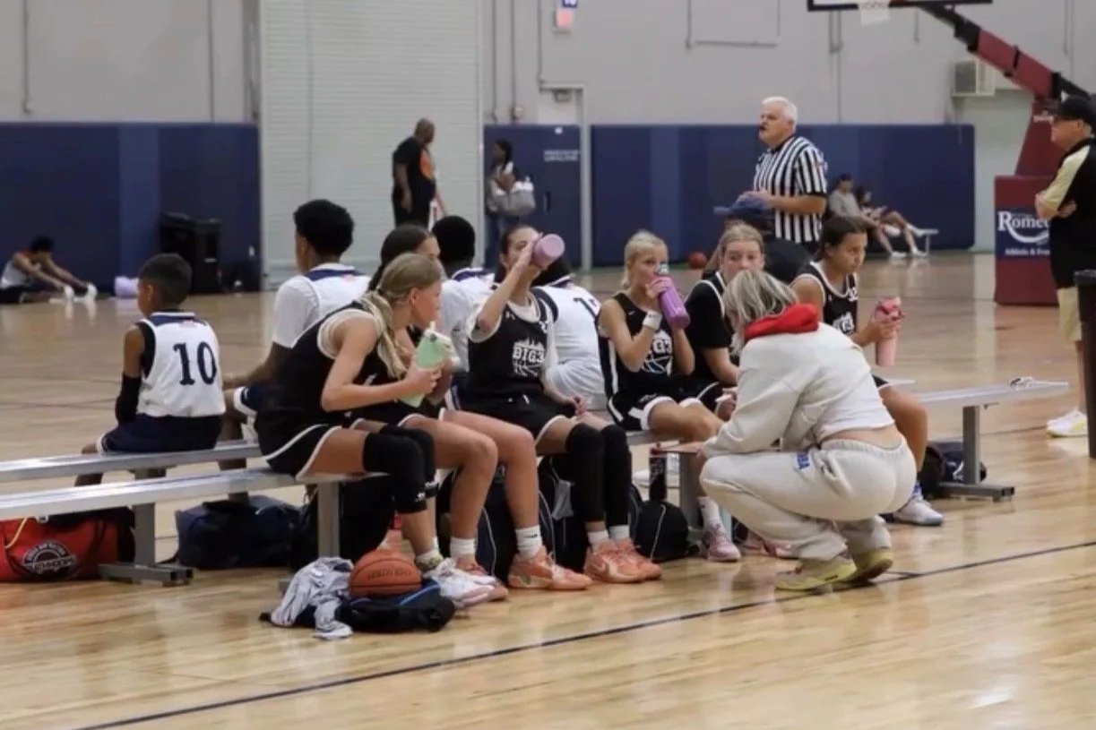 Kids on a basketball team sitting on a bench during a game or practice, with a coach or team assistant kneeling in front of them talking.