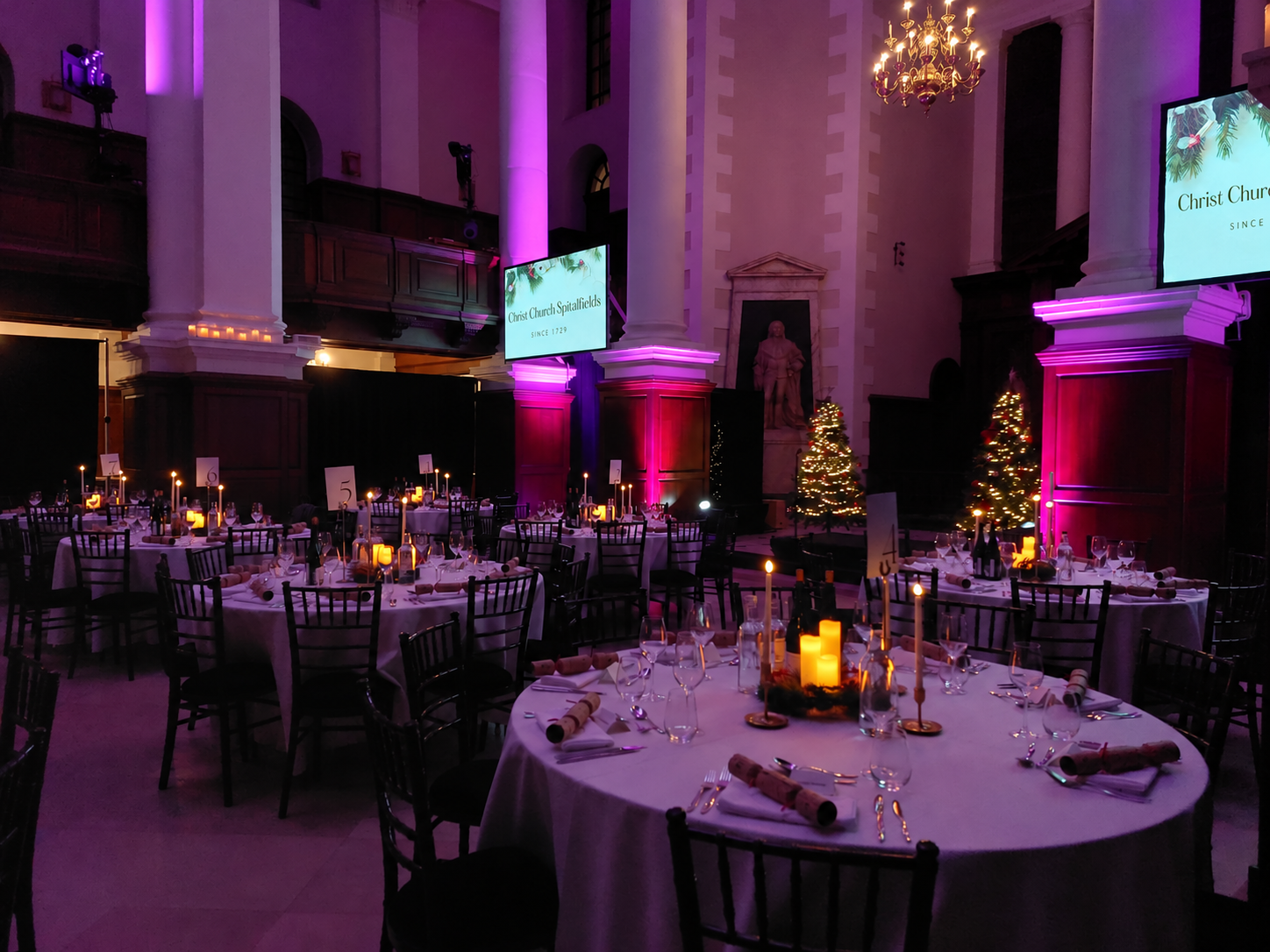 Round tables set for Christmas dinner in the Nave, with decorated trees and purple uplighting