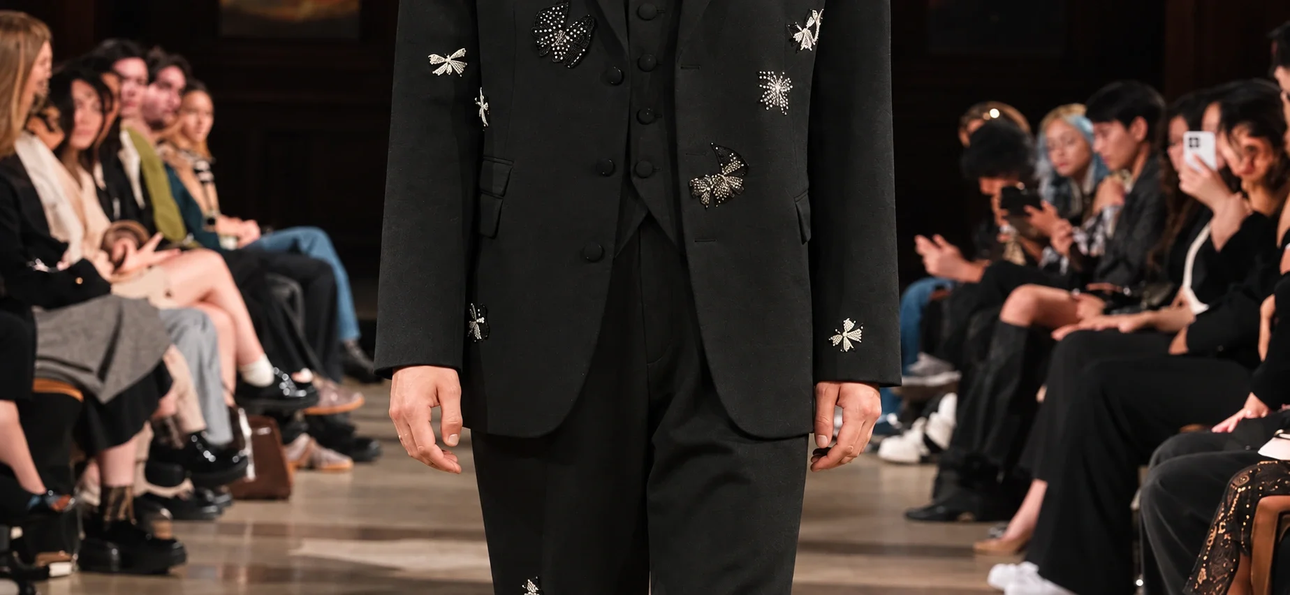 Model wearing a black embellished suit on a runway between seated guests in the Nave