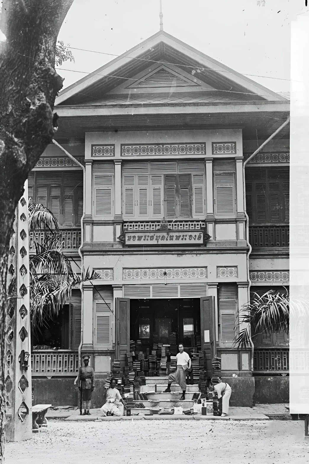 Black and white photo of a traditional Thai building with Thai script on a sign above the entrance. In front, there are five people and stacks of books placed on the stairs and ground, indicating some form of book sale or distribution. There are trees and plants on either side of the building.