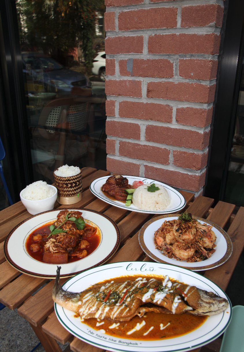 A wooden table outside a restaurant with several plates of food, including rice, a spicy stew with vegetables, fried seafood, a grilled fish in curry sauce, and sliced grilled pork with rice, against a brick wall and glass window reflecting parked cars.