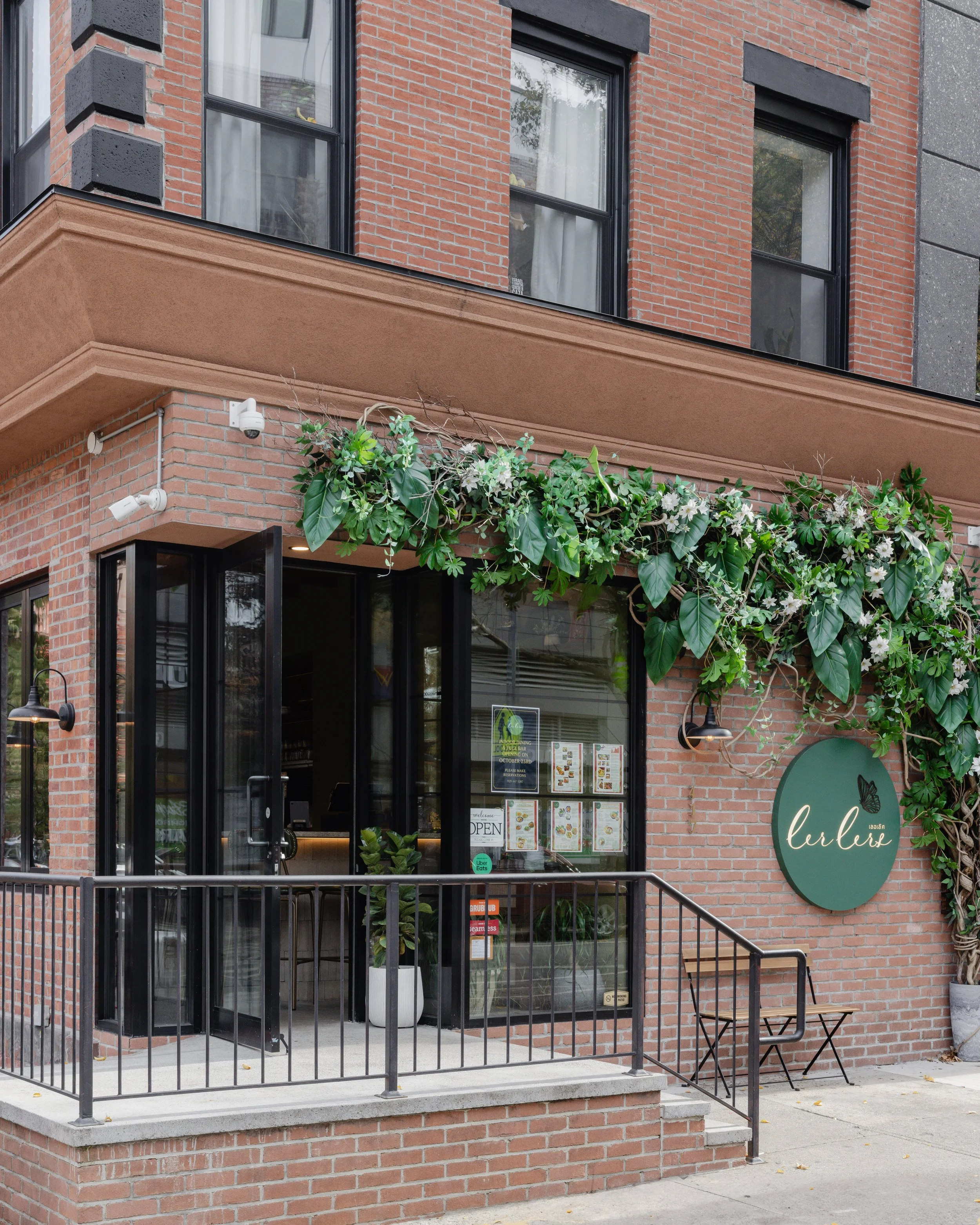 estaurant entrance. The terrace has a black metal railing, black-framed glass doors, and large windows. There are hanging black lamps, a sign that says "lerler" with a butterfly logo, green plants, and a small seating area outside with a chair and a side table.