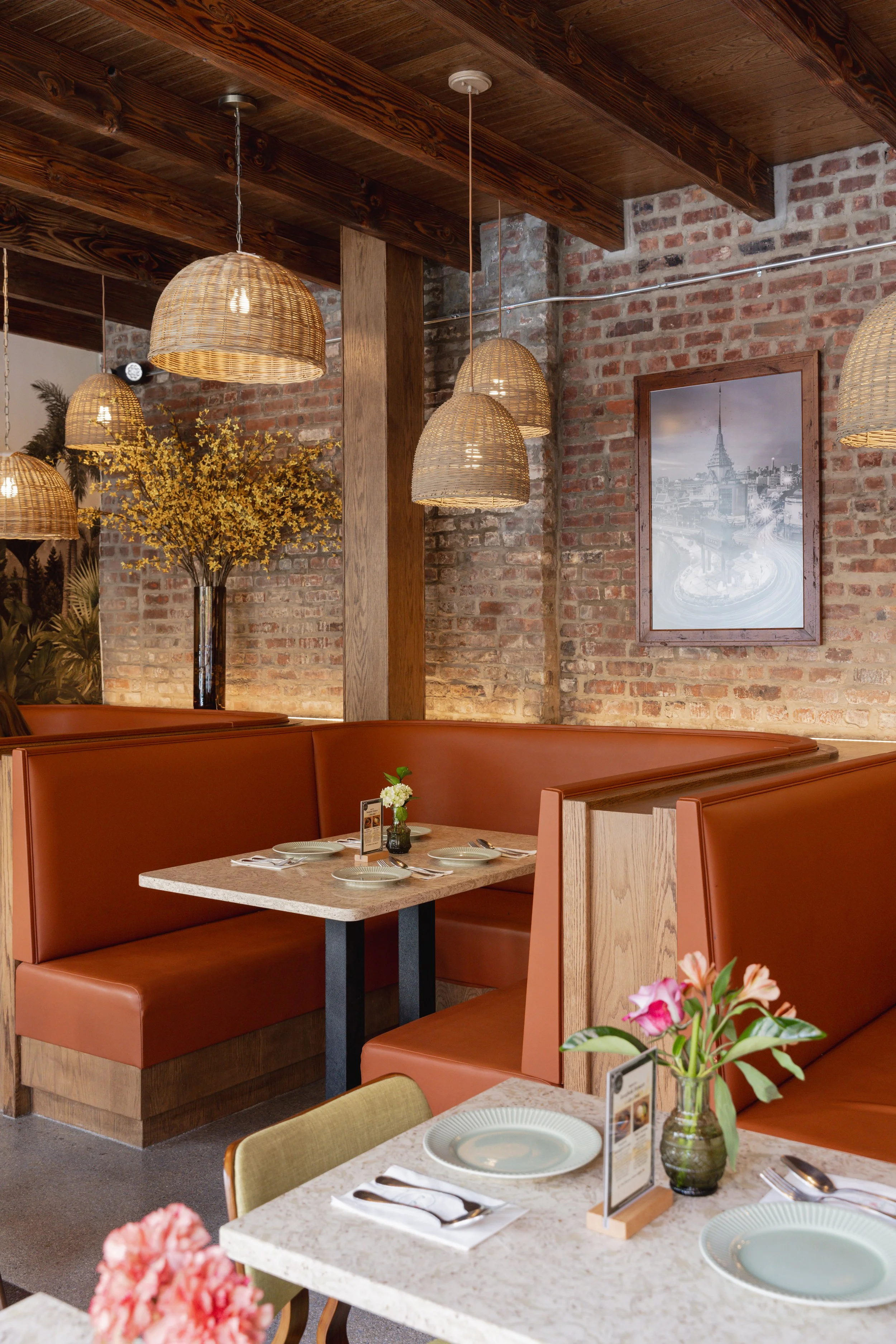 The image shows a cozy restaurant interior with exposed brick walls, wooden beams, hanging wicker pendant lights, and decor including a framed black-and-white cityscape photo and a tall vase with yellow flowers. There are tables set with plates, silverware, and small flower arrangements.