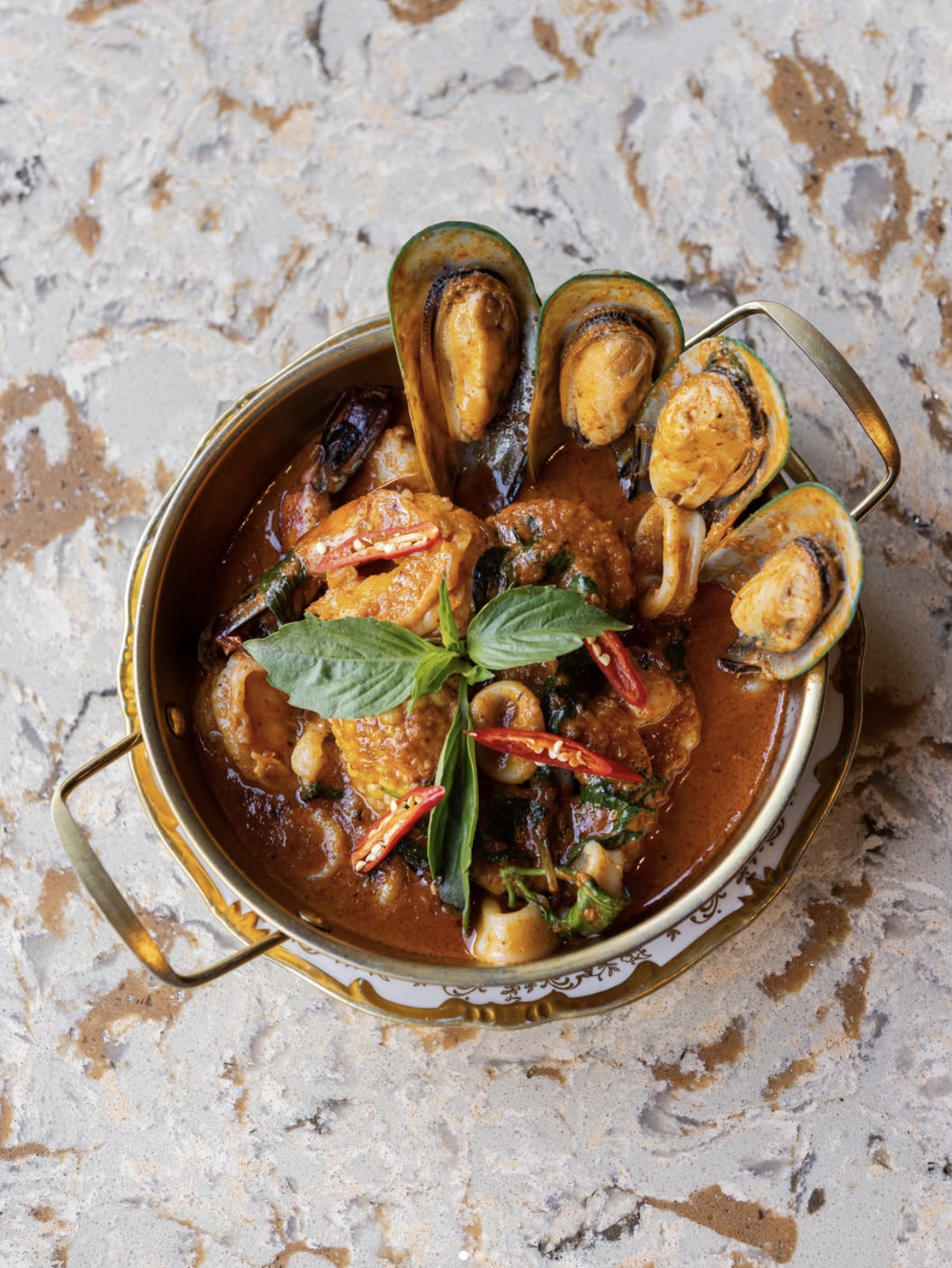 Seafood dish featuring mussels, shrimp, and squid in a red sauce, garnished with basil leaves and red chili slices, served in a silver bowl on a textured surface.
