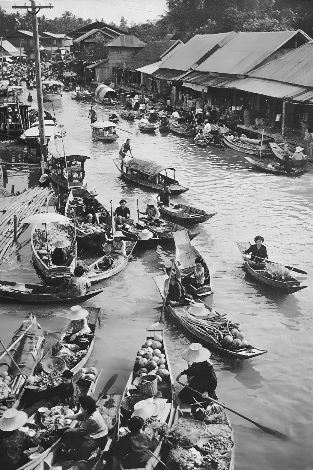 A busy river market scene with numerous small boats filled with vendors and shoppers, along a dockside with buildings in the background.