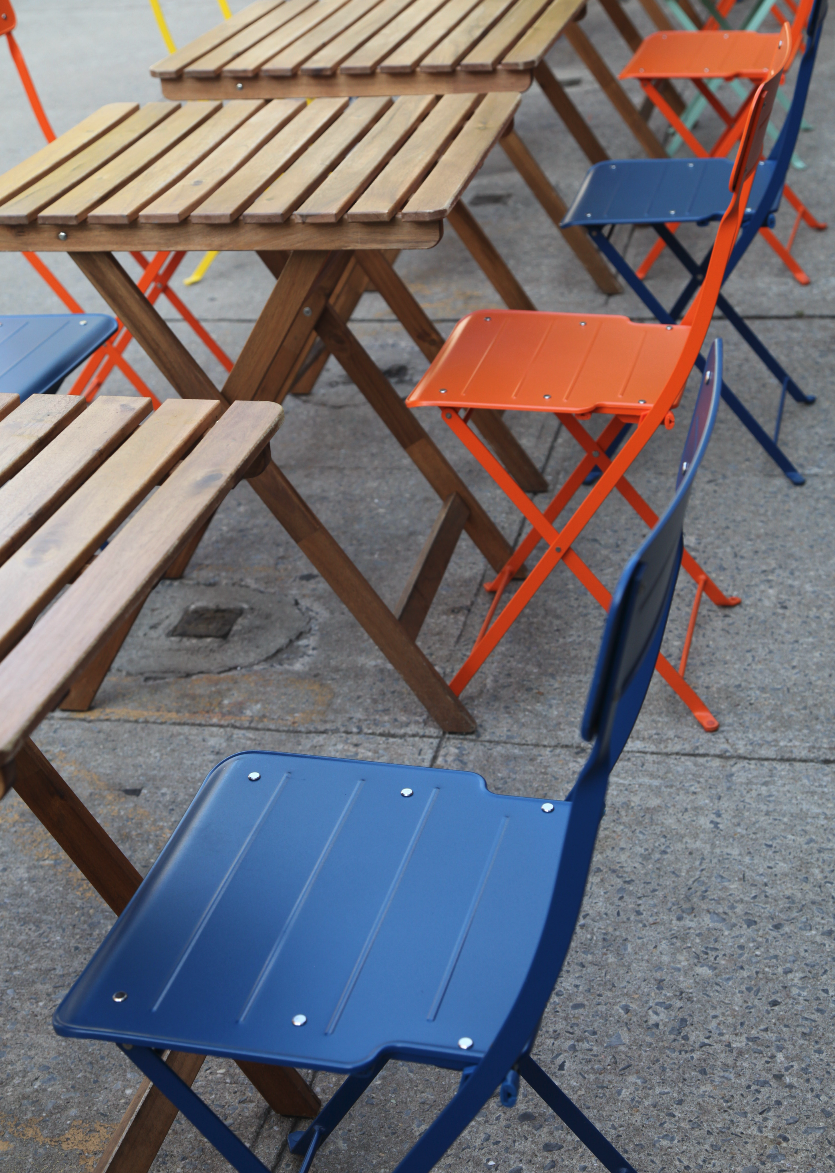 A row of colorful metal and wooden chairs and tables set up outdoors, with the chairs in blue and orange, and the tables made of wooden slats.