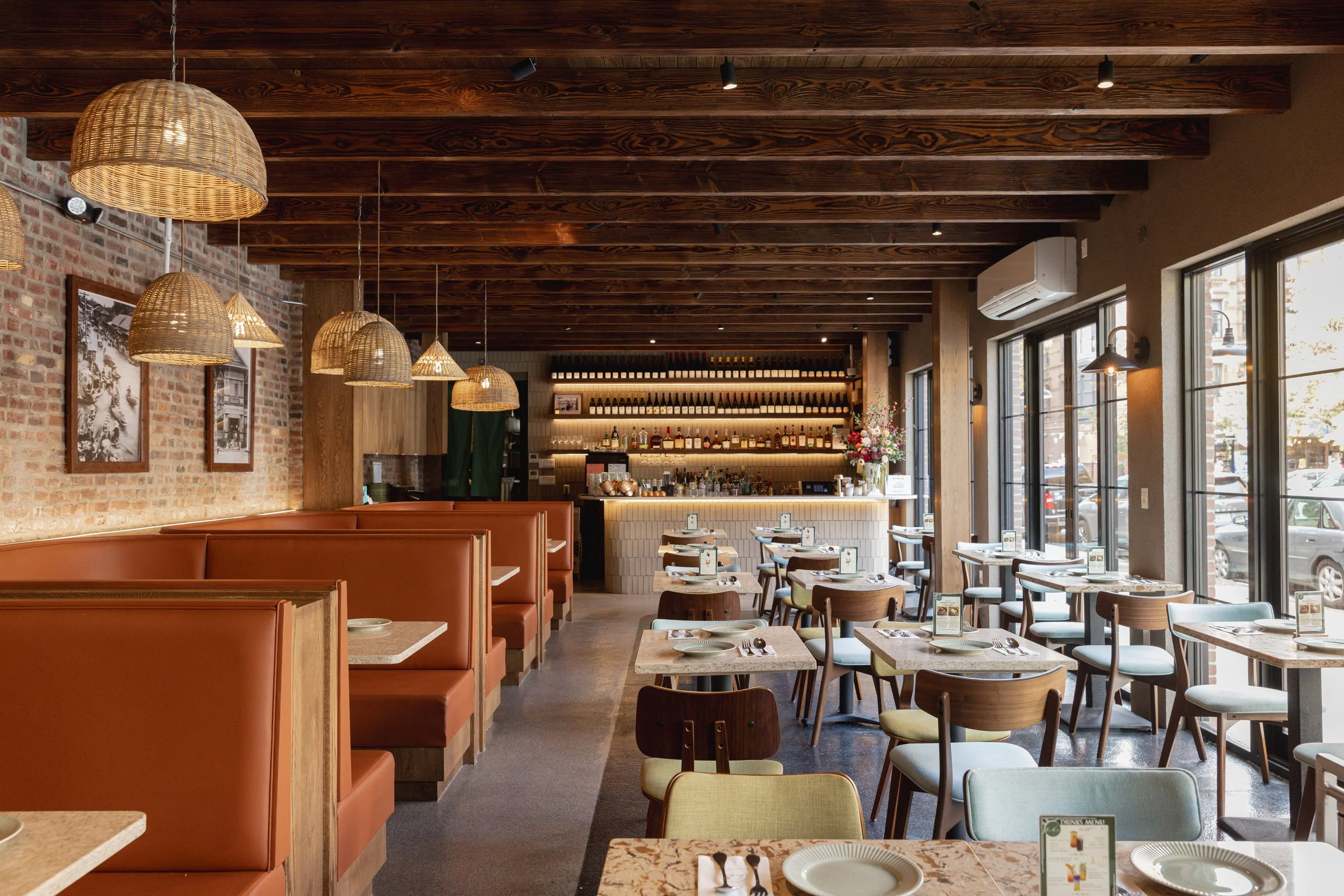 Inside a cozy restaurant with exposed brick walls, wooden ceiling beams, hanging wicker lights, and large windows letting in natural light. There's a bar with wine bottles at the back and several tables set for dining.