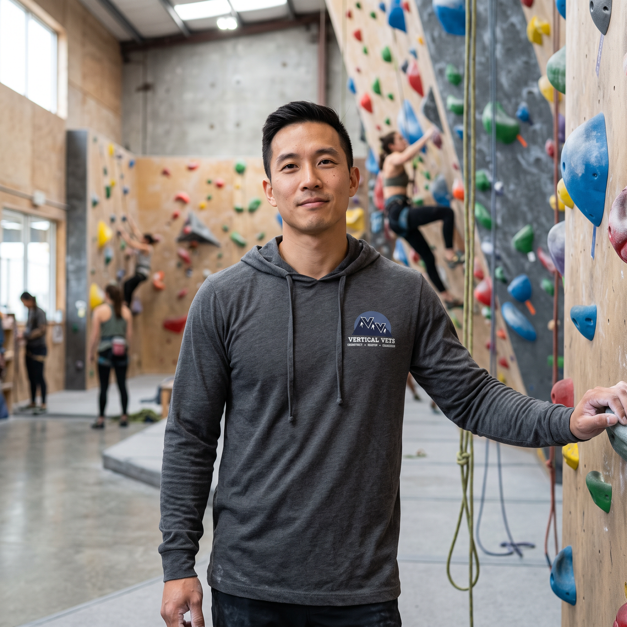 A young man with dark hair and a gray hoodie stands in front of a climbing wall in an indoor rock climbing gym, with other climbers in the background.