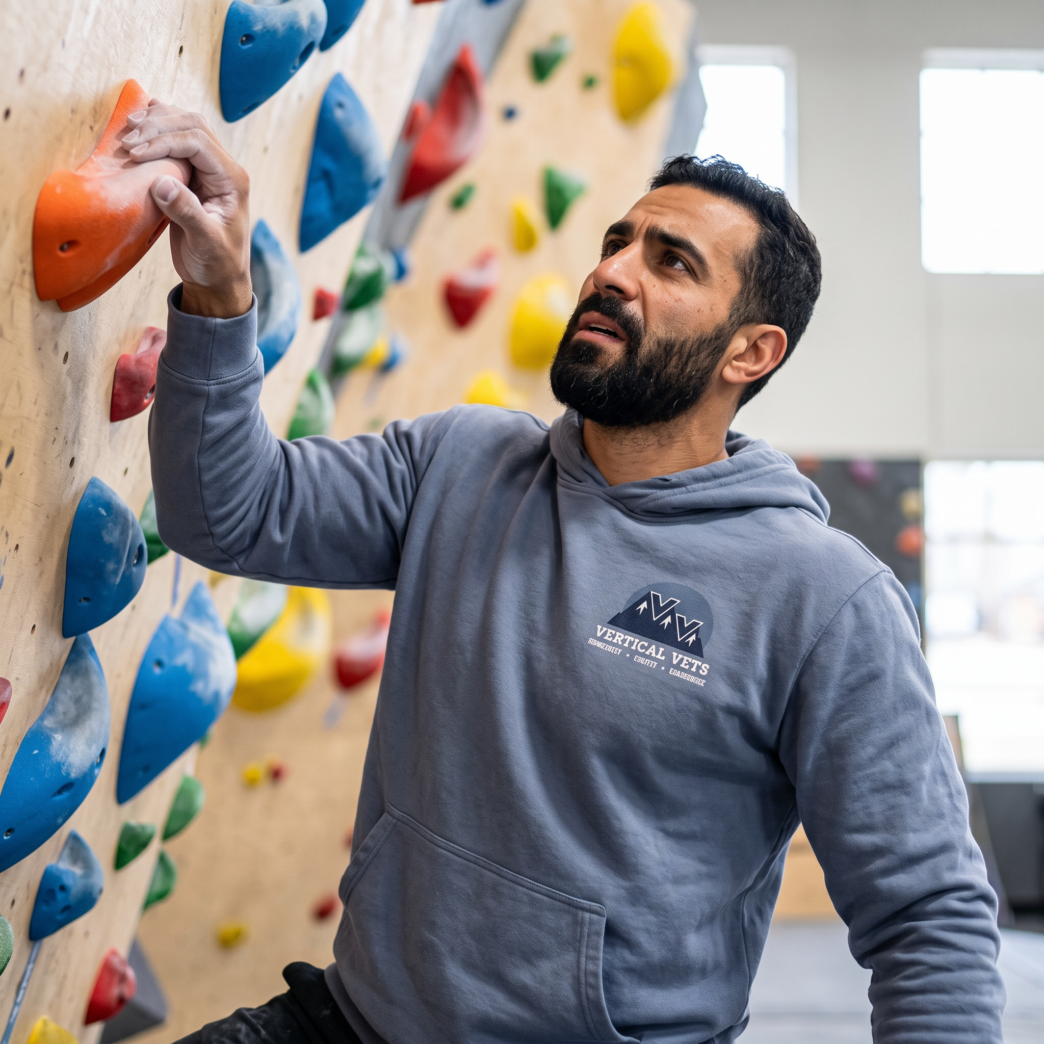 A man bouldering on an indoor climbing wall filled with colorful holds. He is wearing a gray hoodie and has a focused expression.