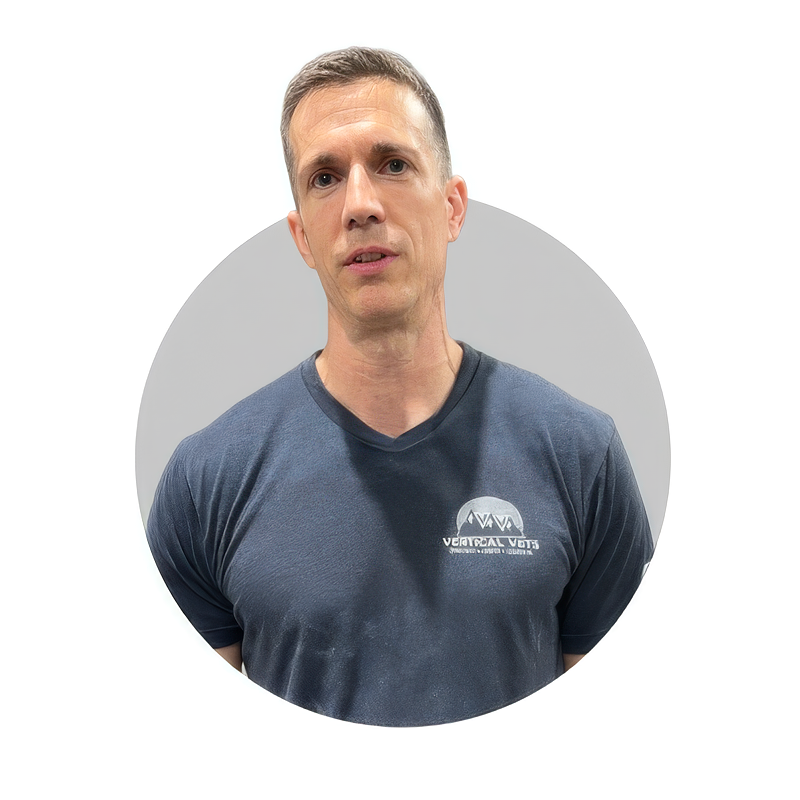 A man with short hair wearing a dark gray T-shirt with a logo on the chest, standing in front of a plain background.