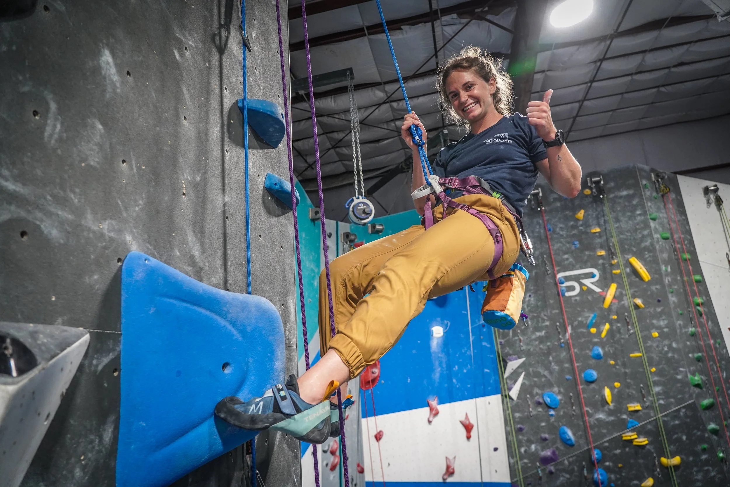 A woman with curly hair wearing climbing gear and a navy T-shirt giving a thumbs up while bouldering at an indoor climbing gym.