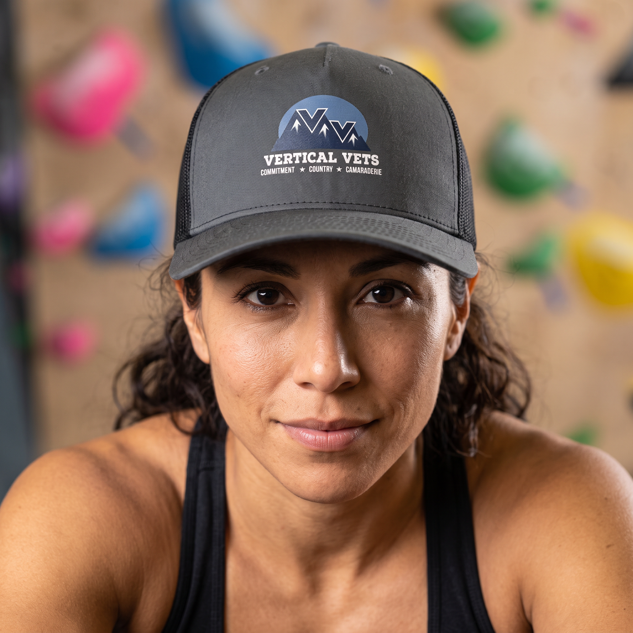 A person wearing a gray baseball cap with the logo 'Vertical Vets' and mountain graphics, looking upward at a climbing wall with colorful holds.