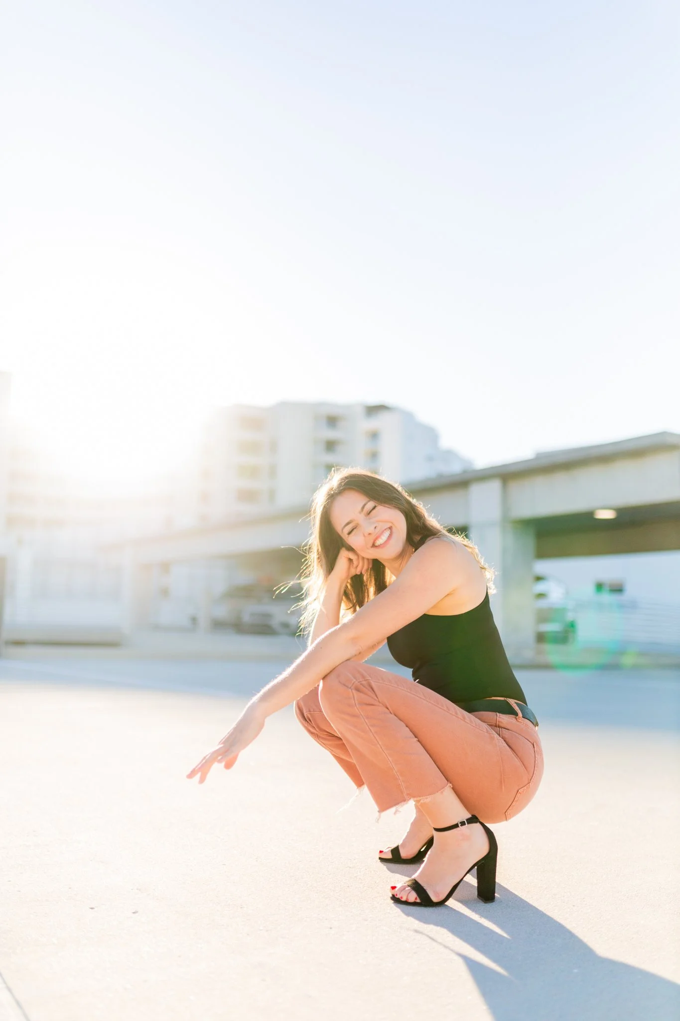 A woman, Lindsey Tosoni, with long brown hair squatting on a city parking lot, smiling at the camera, wearing a black sleeveless top, pink pants, and black high heels, with buildings and a bridge in the background and sunlight shining from the left.