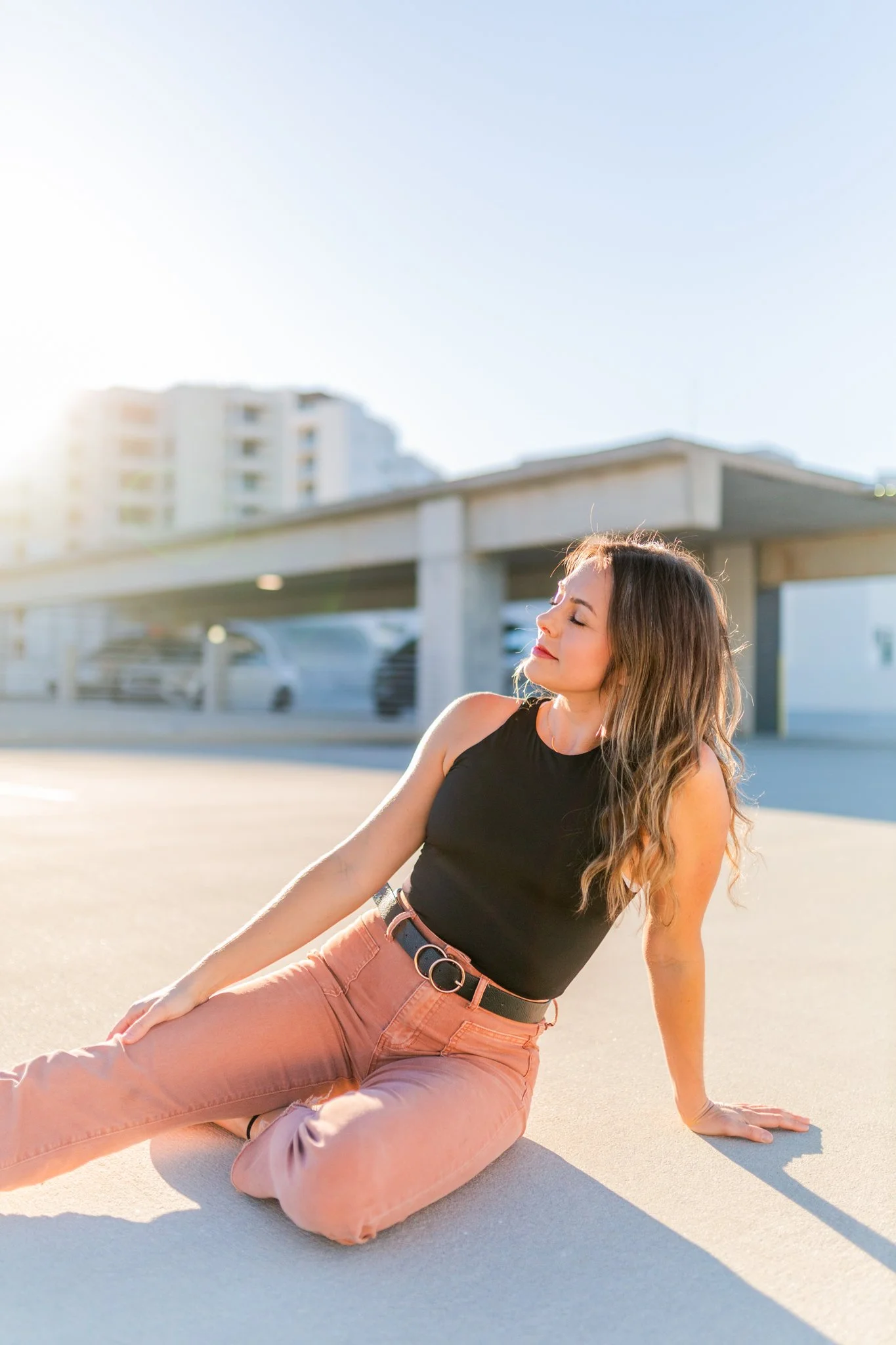 Young woman, Lindsey Tosoni, with long wavy hair wearing a black sleeveless top and pink pants, sitting on the ground outdoors in sunlight with a building and parking garage in the background.