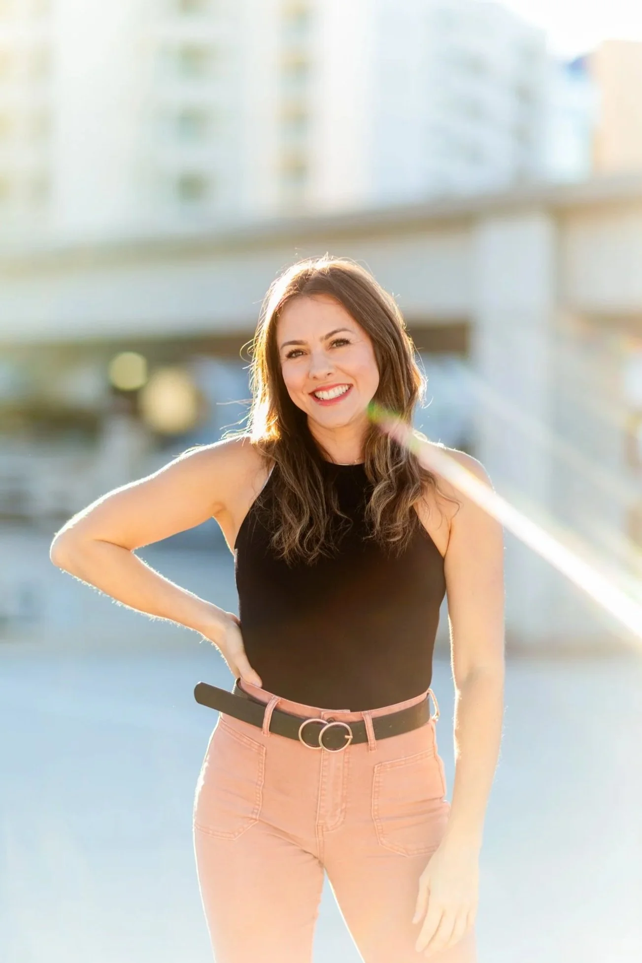 A woman, Lindsey Tosoni, smiling outdoors during the daytime with her hand on her hip, wearing a black sleeveless top and light pink pants, with a cityscape background.