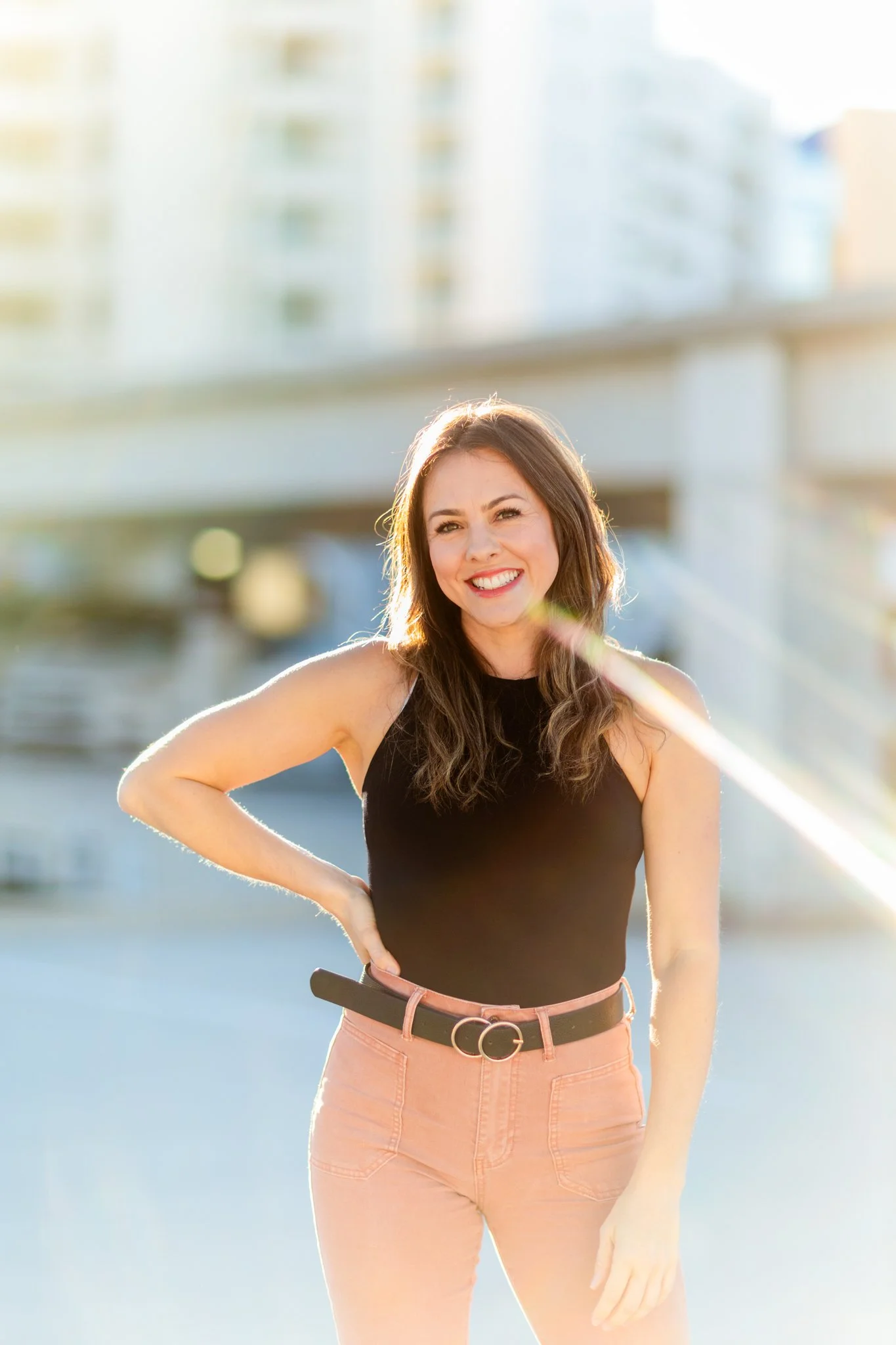 A woman, Lindsey Tosoni, smiling outdoors with her hand on her hip, standing near a cityscape in the background, wearing a black sleeveless top and pink pants.